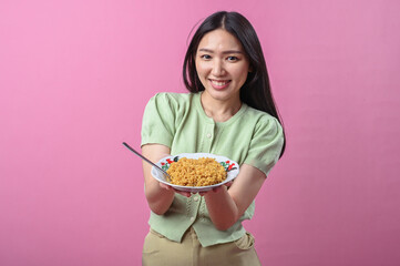 Asian woman smiling while holding a plate of cooked instant noodles with a spoon, wearing a green...
