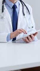 Adult male doctor wearing lab coat and stethoscope using digital tablet while sitting at desk in medical office, close up and vertical view. Medicine, health care concept