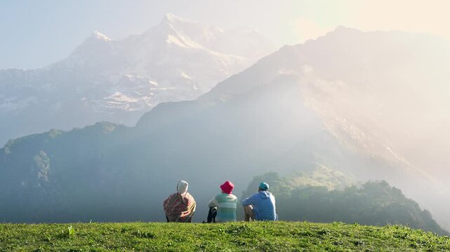 Young men sit on the edge of a mountain, taking in the view of the valley with trees, blue sky, and surrounding mountains along the Madmaheshwar Temple trek, near Ransi Village, Uttarakhand, India.