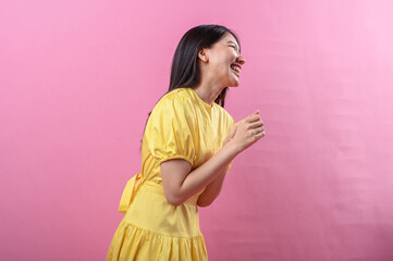 Asian woman in a yellow dress laughing joyfully while facing sideways against a soft pink...