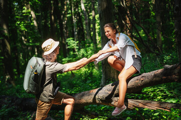 Young male traveler helping girl to climb over fallen tree during trekking in summer forest