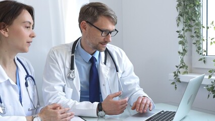 Two doctors are studying medical charts on a laptop, discussing patient data in a hospital or...
