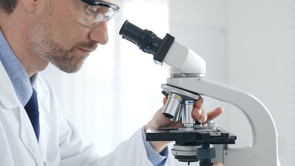 Adult male scientific researcher analyzing sample through microscope, wearing protective gear in...