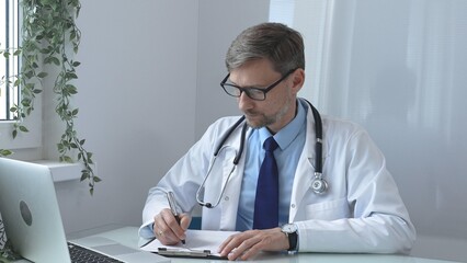 Doctor man in glasses documents patient information, takes notes on clipboard, sitting at table...