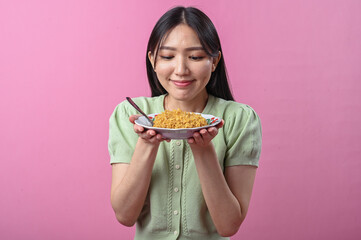 Asian woman in green top laughing joyfully while holding a forkful of instant noodles above a plate, in front of a vibrant pink background