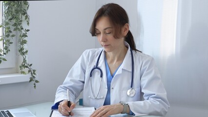Female doctor in lab coat and stethoscope diligently writing notes on clipboard while sitting at...