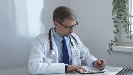 Doctor man documenting patient information, writing notes on clipboard while seated at desk, laptop...