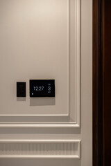 Close-up of a smart home control panel displaying the time, day, and date (12:27, Tue, 3 Oct), and a light switch on a white paneled wall.