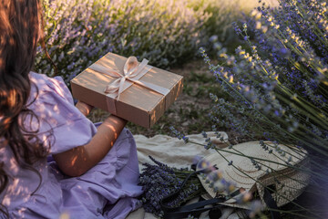 Girl in lavender dress holding a gift box with ribbon, sitting among blooming lavender flowers....