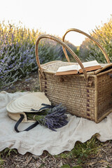 Wicker picnic basket, open book, lavender bouquet, and straw hat on blanket in blooming lavender...