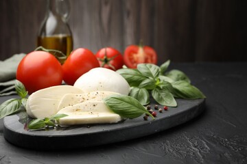 Tasty mozzarella cheese, tomatoes, basil, peppercorns and oil on black table, closeup