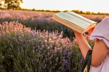 Girl in lavender field reading book at sunset. Holding a straw bag with lavender bouquet, dressed...