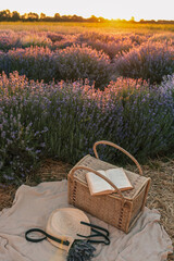 Wicker picnic basket with open book, lavender bouquet, and straw hat on blanket in lavender field...