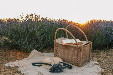 Wicker picnic basket with open book, lavender bouquet, and straw hat on blanket in lavender field...