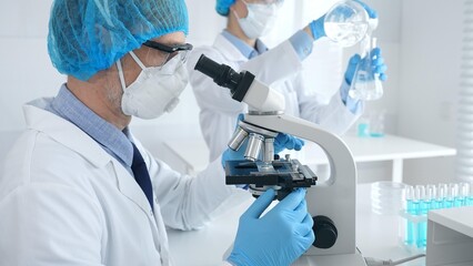 Masked male scientist examining specimen under microscope while laboratory assistant handles...