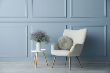 Comfortable armchair and vase with flowers near light blue wall indoors