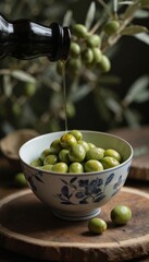 Green olives in a white bowl on wooden board. Olive oil bottle poured over olives in a kitchen setting. Fresh produce, natural light. Wooden board, olive tree blurred in the background.