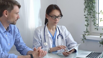 Female doctor using a digital tablet for explaining test results to a male patient during a consultation in a bright, modern medical office. Medicine concept