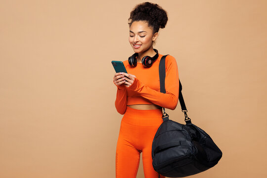Young fitness trainer instructor sporty woman sportsman she wear orange clothes spend time in home gym hold bag use mobile cell phone isolated on plain beige background. Workout sport fit abs concept.