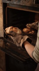 Hands of baker pulling freshly baked bread out of oven with oven mitts in rustic kitchen setting.