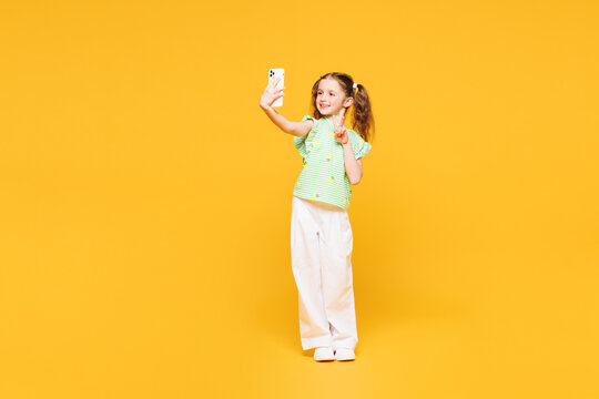 Full body little cute kid child girl 8-9 years old wear green t-shirt doing selfie shot on mobile cell phone show v-sign isolated on plain yellow background Mother's Day love family lifestyle concept