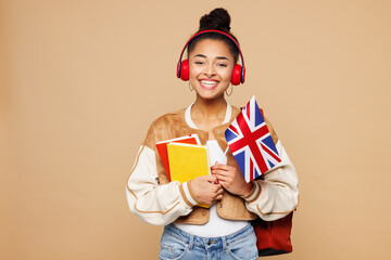 Young smart girl student wear casual clothes backpack bag hold books British flag listen to music in headphones isolated on plain pastel light beige background. High school university college concept.
