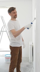 Middle aged man wearing white t-shirt and protective gloves, is painting white wall with paint roller, experiencing home renovation and transforming living space, vertical view