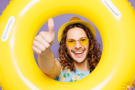 Close up young man wear blue beach t-shirt yellow hat sunglasses rest near hotel pool look through rubber ring show thumb up isolated on plain purple background. Summer vacation sea sun tan concept.