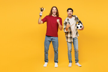 Fototapeta premium Full body young winner two friends men sport fans wears casual clothes cheer up support football team hold soccer ball golden cup watch tv live stream isolated on yellow background. Leisure concept.