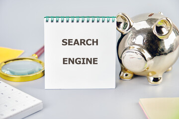A notepad displaying the term search engine is placed alongside a golden piggy bank and a magnifying glass, symbolizing business innovation