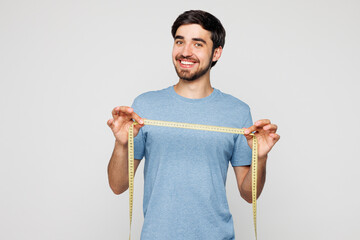 Young smiling happy man wear blue t-shirt casual clothes hold measuring tape show result look camera isolated on plain solid white background. Proper nutrition healthy food unhealthy choice concept.