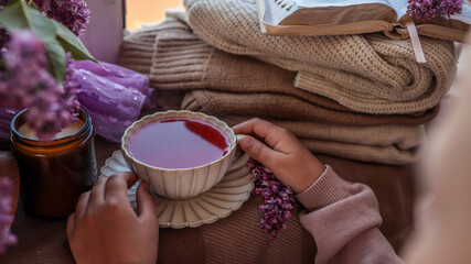 Cozy scene with a cup of berry tea held in hands, lilac flowers, folded knit sweaters, an open...