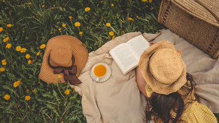 Girl in straw hat reading a book on a picnic blanket among dandelions, with a cup of tea, woven...