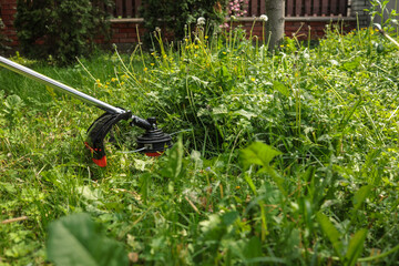 Close-up of grass trimmer cutting overgrown weeds and dandelions in backyard. Concept of garden...