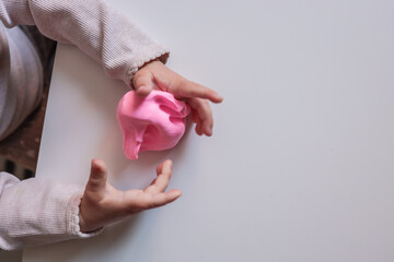 Small child squeezing soft pink modeling clay on a white table. Close-up of hands engaged in...