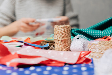Close-up of jute twine, lace, and colorful fabrics on a crafting table with a blurred person working in the background. Creative DIY workspace with handmade decoration materials.