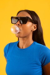 Stylish model blowing bubblegum while wearing sunglasses on yellow background