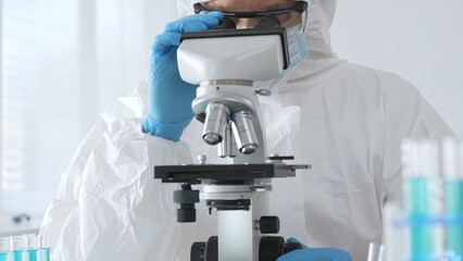 A male doctor in a protective suit and gloves uses a microscope while analyzing samples, conducting...
