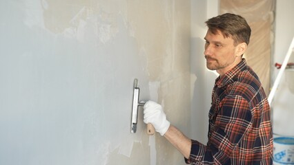 Male construction worker wearing protective gloves smoothing plaster onto wall surface during home...