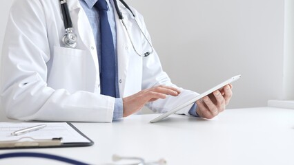 Doctor man wearing white medical coat and stethoscope using digital tablet while sitting at desk in...