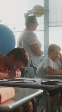 Elementary School Students Writing Exam or Doing Tasks in Notebooks. Female Teacher Walking Between Desks, Controlling Young Boys and Girls During School Test in Classroom. Dolly Shot. Vertical Shot.