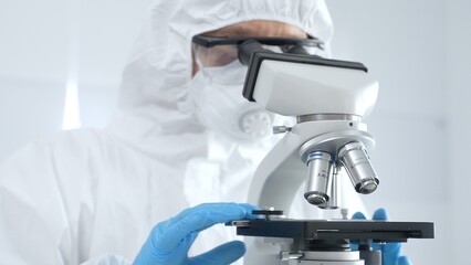 Medical virologist in a hazmat suit, mask, and blue protective gloves, carefully working at a microscope in a laboratory researching viruses. Healthcare and science concept