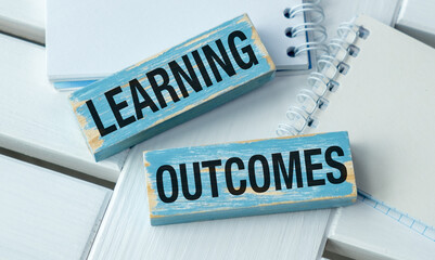 Two wooden blocks with the word Learning Outcomes on them. One block is on top of the other. There is a spiral notebook on the table