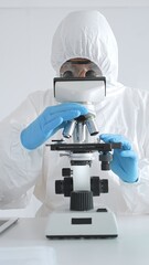 A medical virologist in protective gear examines viral samples under a microscope while conducting...