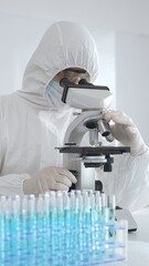 A medical virologist in protective gear examines viral samples under a microscope while conducting...