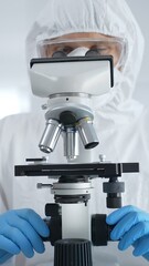 Male virologist in a hazmat suit, mask, and blue gloves, diligently working at a microscope in a...