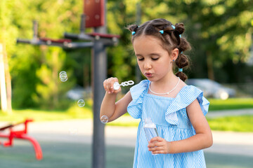 Girl in a blue striped dress concentrates while blowing several soap bubbles from a wand at a leafy...