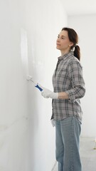 Middle aged happy woman wearing checked shirt, protective gloves, is rolling white paint onto wall,...