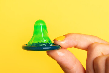 Close-up of a green lubricated condom held between two fingers with yellow nail polish against a...