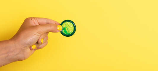  Hand with yellow nails holding a green rolled condom against a bright yellow background, promoting...